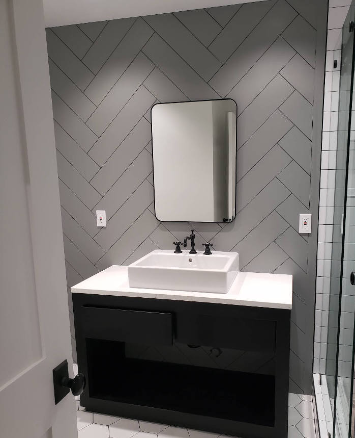 Modern bathroom vanity with gray herringbone tile, white sink, mirror, and black cabinet.