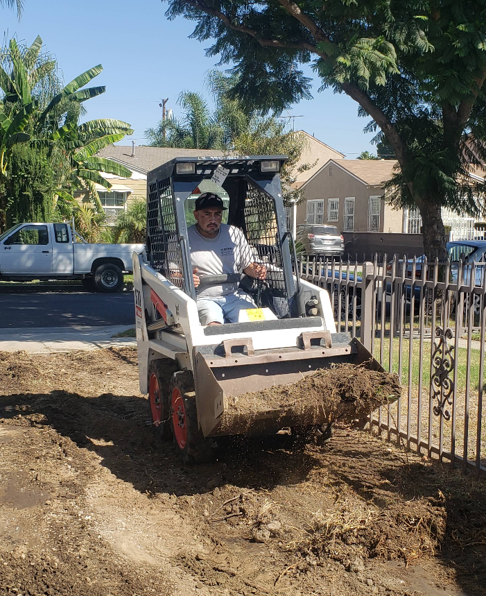 Worker operating a skid steer, pushing dirt beside a fence in a sunny yard