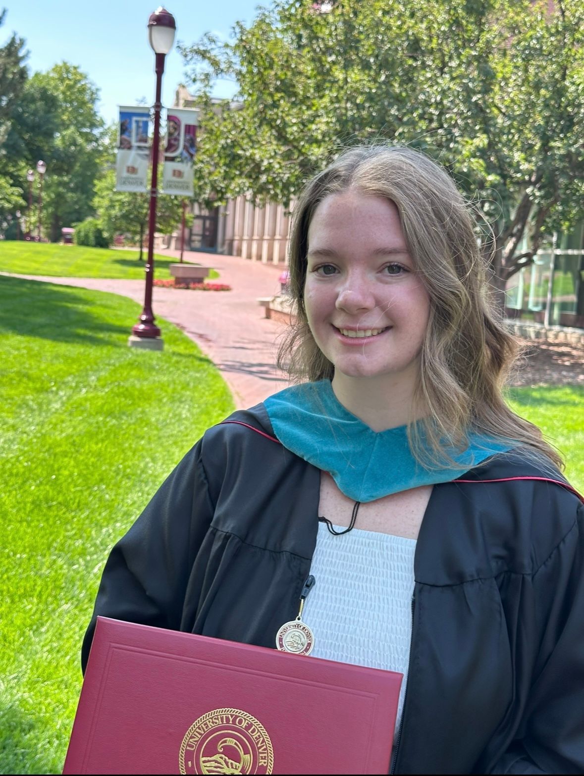A person wearing a black graduation robe, with the teal Master's Degree hood, holding a diploma frame on a sunny day with green grass in the background. 