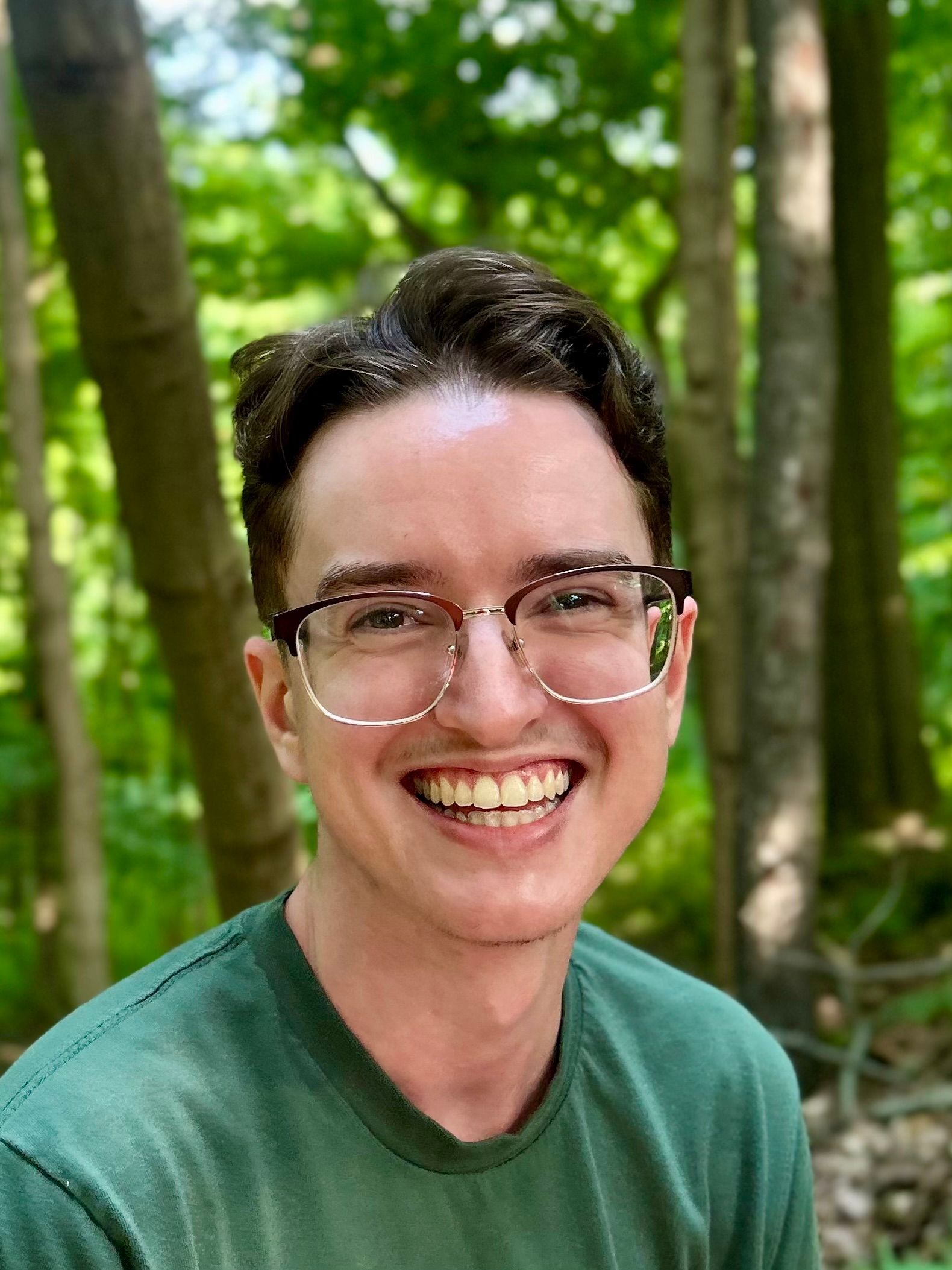 A person wearing glasses and smiling outdoors with trees in the background. 