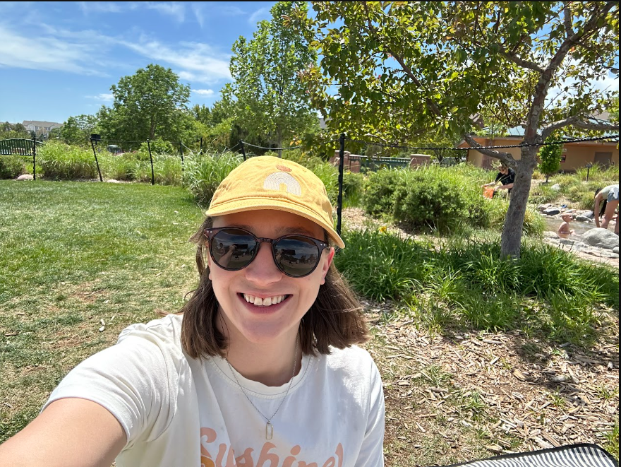 Image of  a person wearing sunglasses and a yellow hat on a sunny day with green trees and grass in the background.