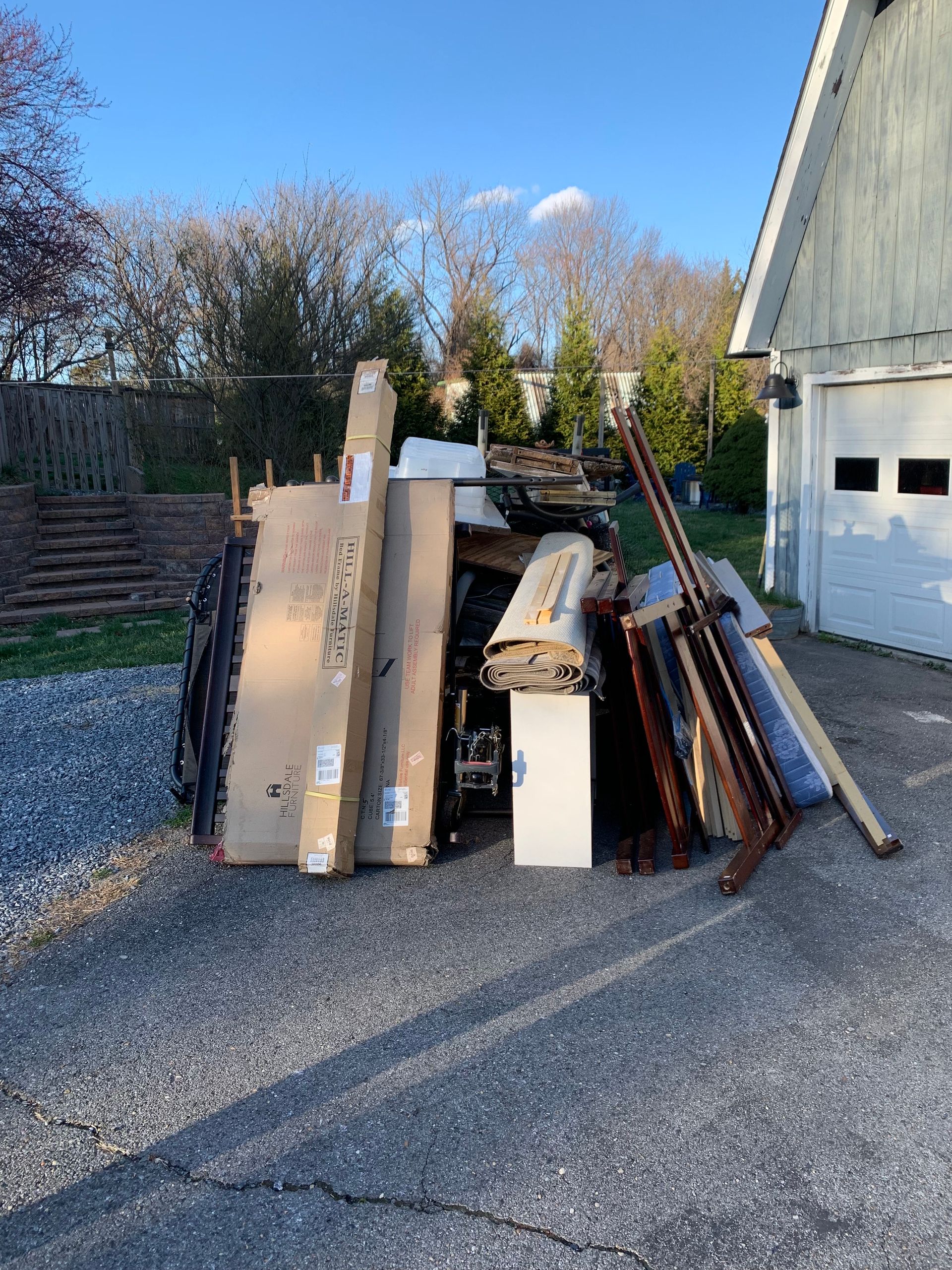 Pile of discarded furniture and appliances on a grassy curb, including a refrigerator, bed frame, and shelving unit.