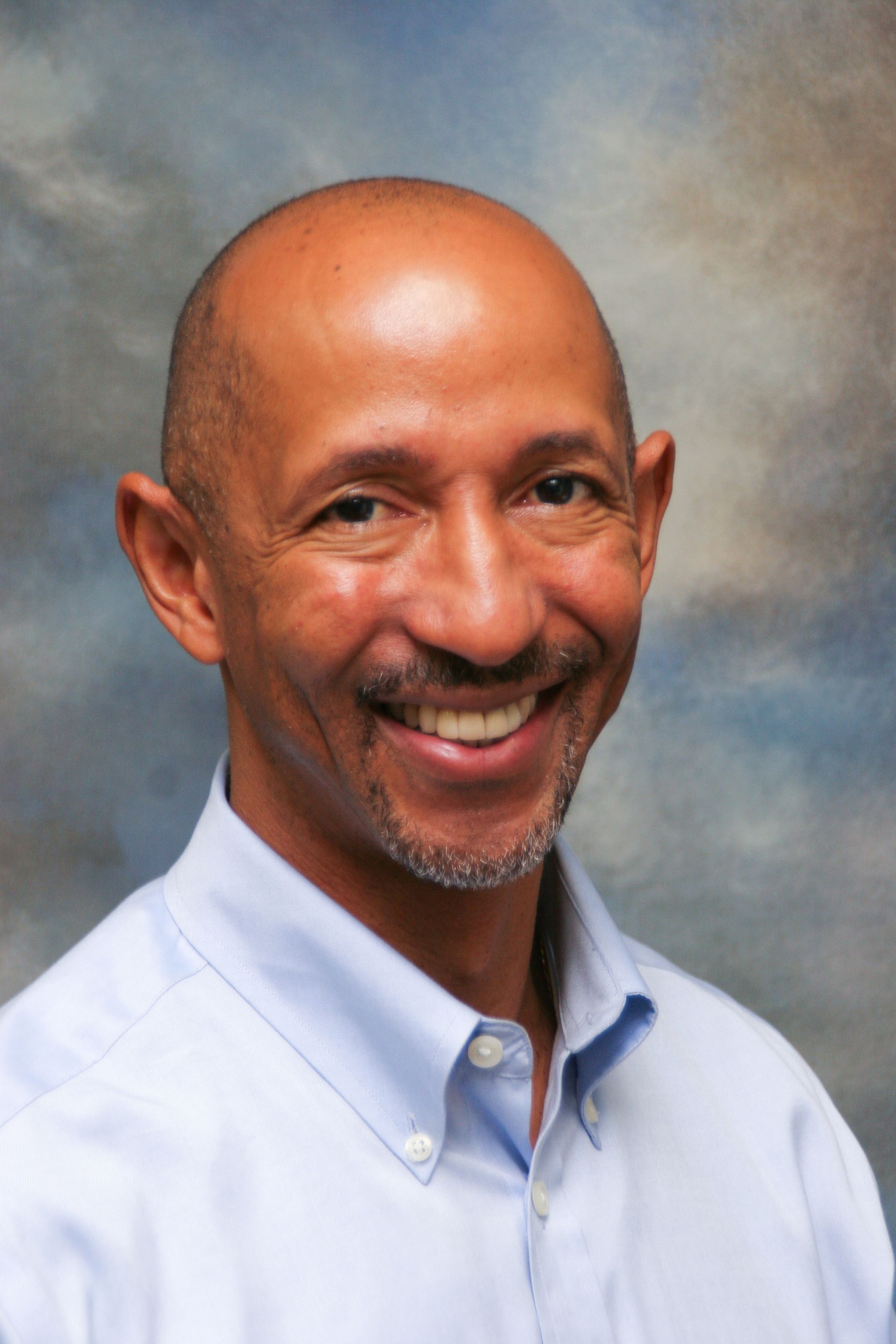 Bald man smiling in light blue shirt, against a soft-focus background.