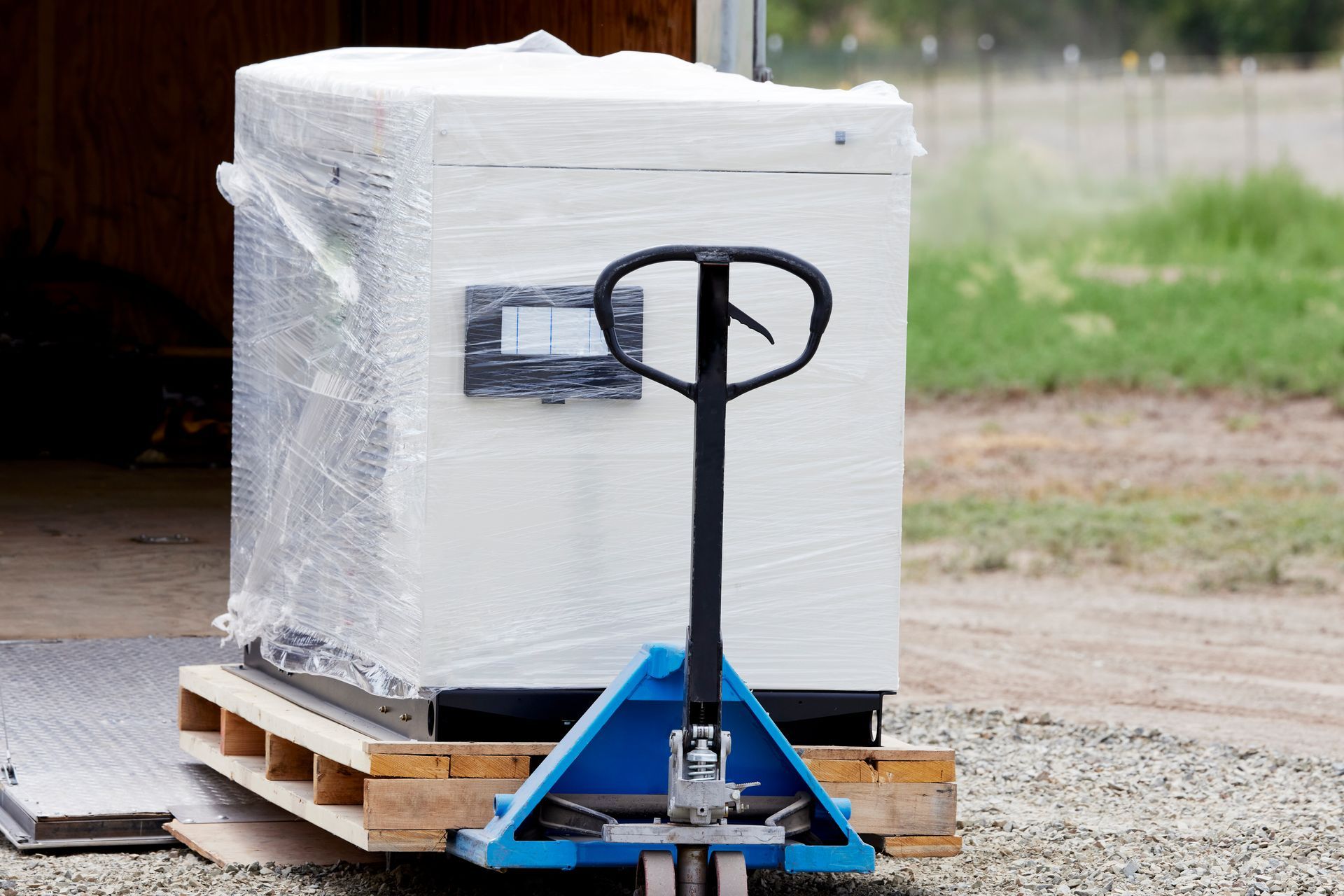 Pallet jack lifting a shrink-wrapped white crate outdoors on a wooden pallet.