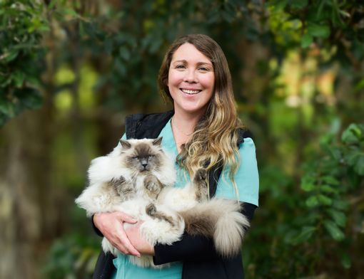 Person holding a fluffy white-and-gray cat outdoors, smiling in front of greenery.