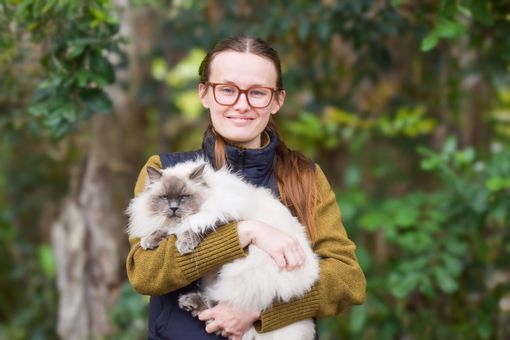 Person holding a fluffy white-and-gray cat outdoors, smiling in front of greenery.