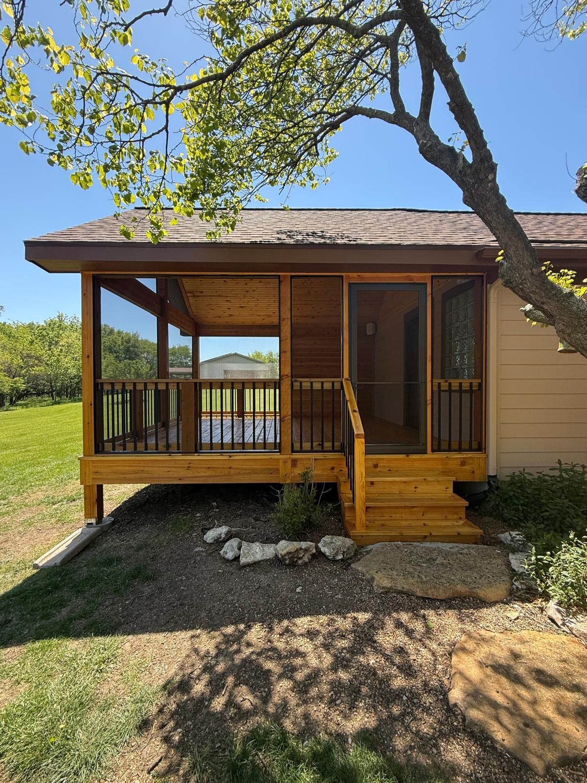 Small wooden porch with steps and railing attached to a beige house under a leafy tree