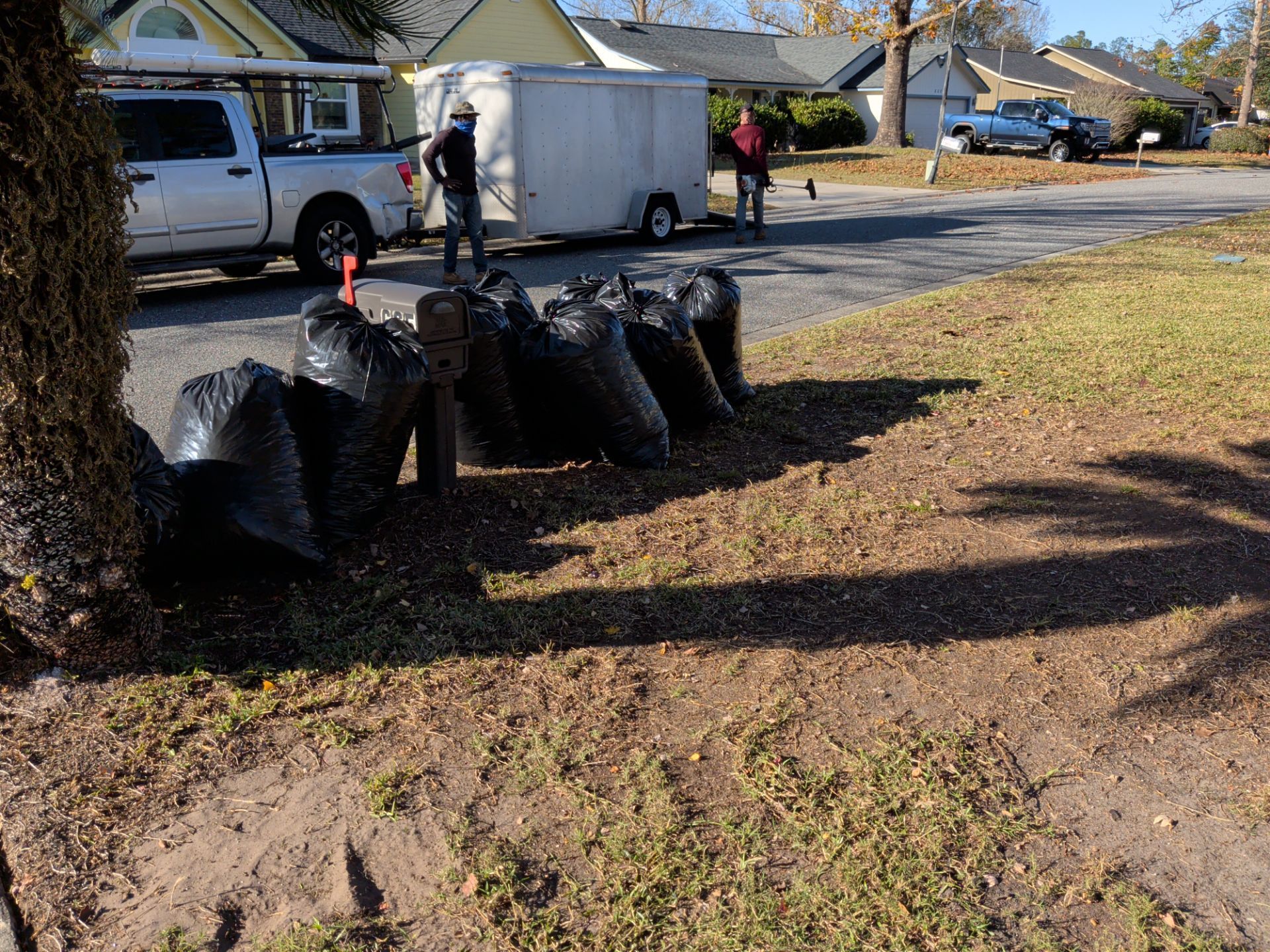 Black trash bags line a yard near a road, with workers by a truck and trailer in the background.