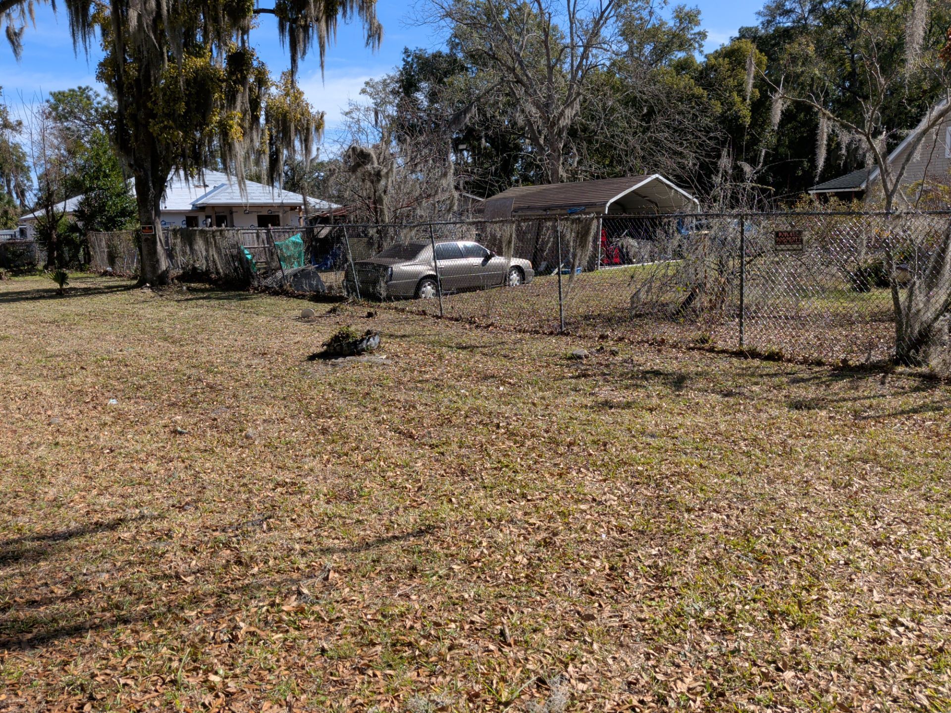 Grassy yard with a pickup truck under a carport. A house and trees are in the background, with a chain-link fence.