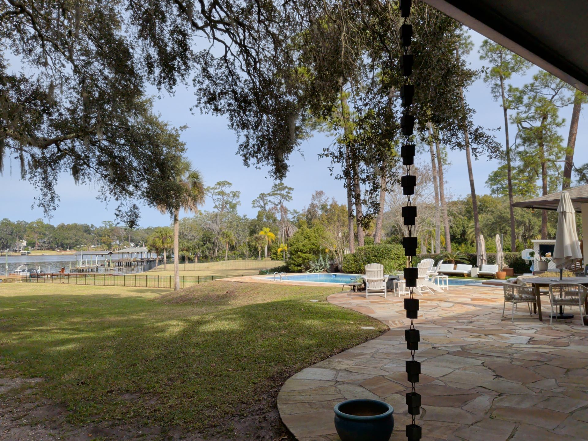 Lush green yard with pool, trees, and waterway in the distance; patio with furniture in the foreground.