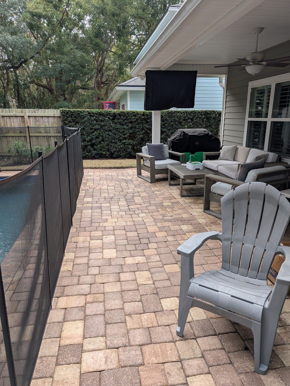 Patio with a pool safety fence. TV mounted on the ceiling, seating area with gray furniture, and brick pavers.