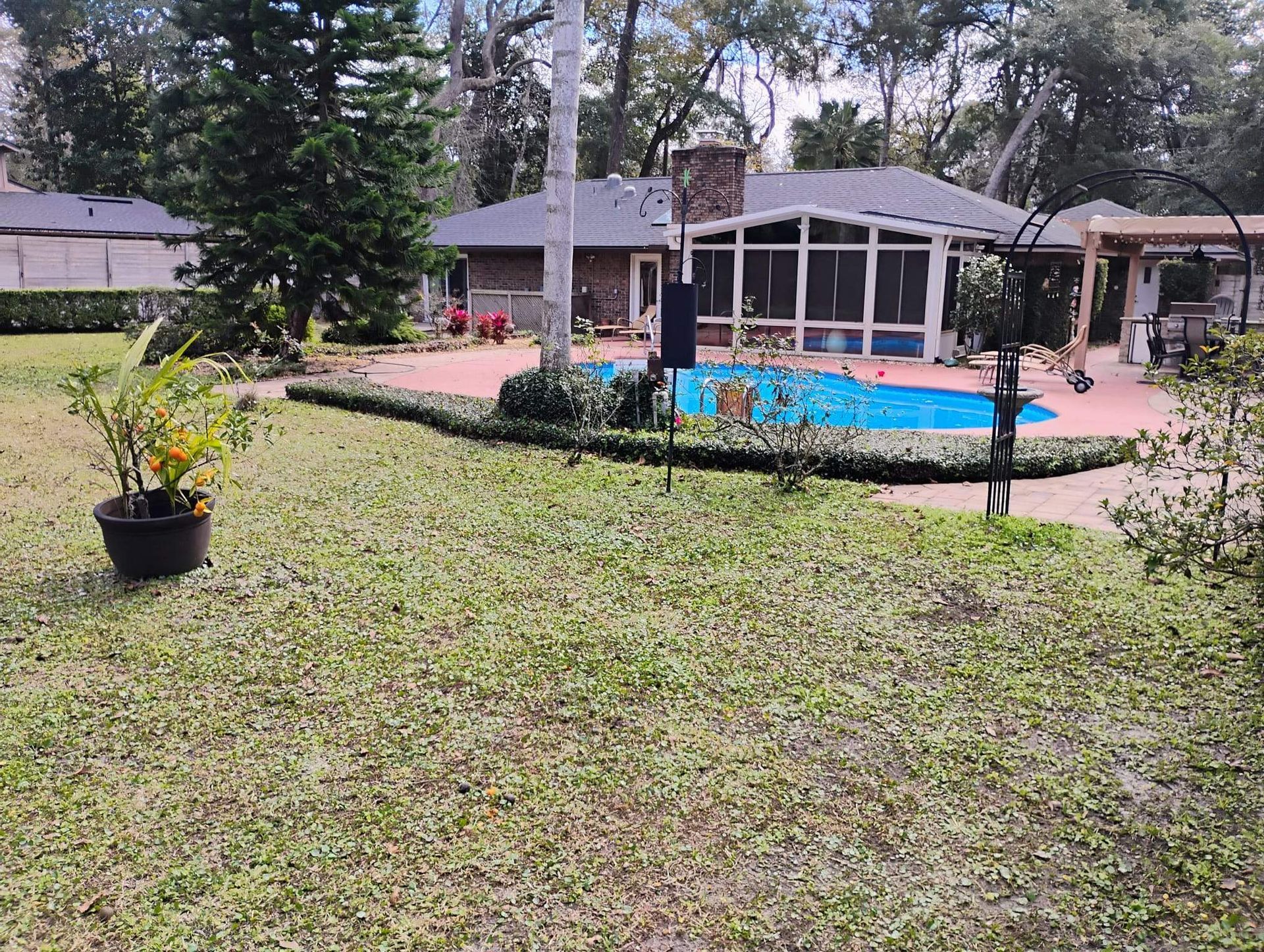 Lawn with potted plant, pool, brick patio, and house with screened porch. Trees in background.