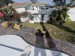 Poolside scene with a house in the background. Grass, patio, and debris present.