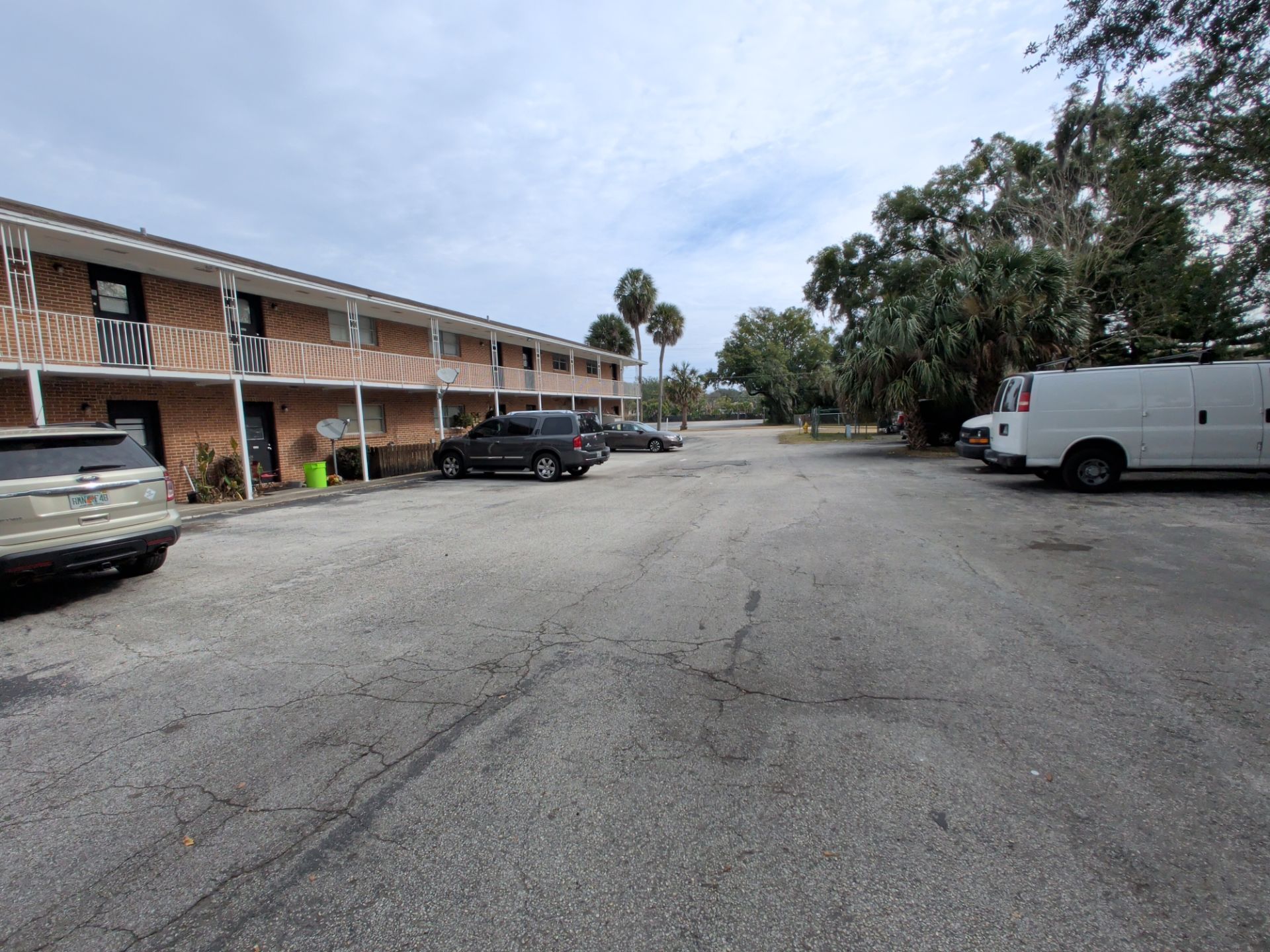 Apartment building with cars parked in front on a cracked asphalt lot, under a cloudy sky.