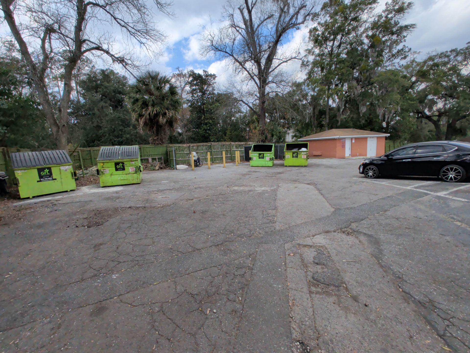 An outdoor parking lot with green dumpsters and a black car. A restroom building is in the background.