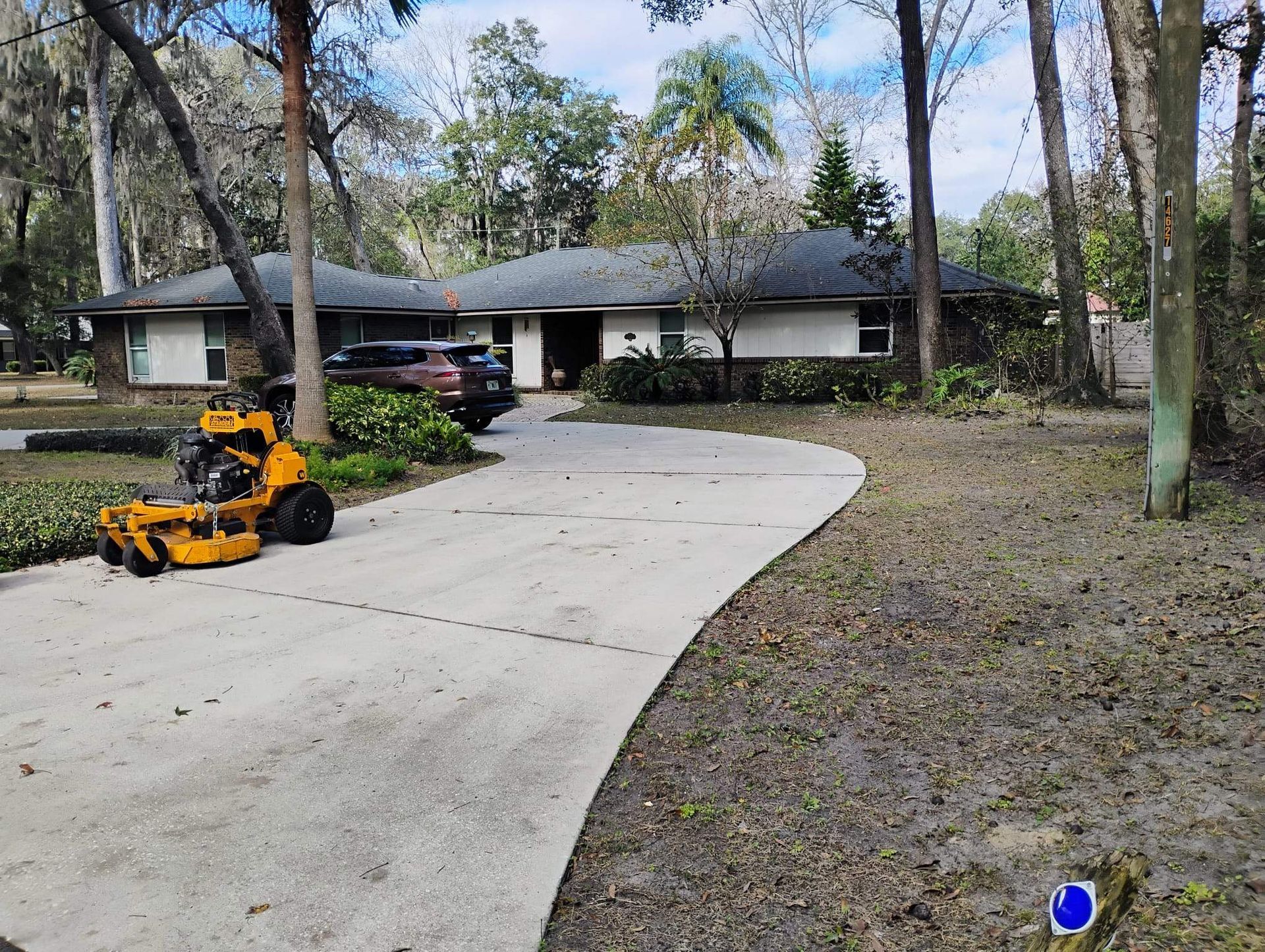 A house with a curved driveway, lawnmower, and a parked car. Trees and foliage surround the property.
