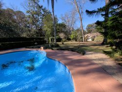 A blue swimming pool curves towards a grassy yard with trees and a small building in the distance.