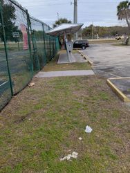A bus shelter along a grassy area next to a chain-link fence and parking lot.