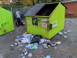 A lime green dumpster overflowing with trash on a paved area, with a person standing nearby.
