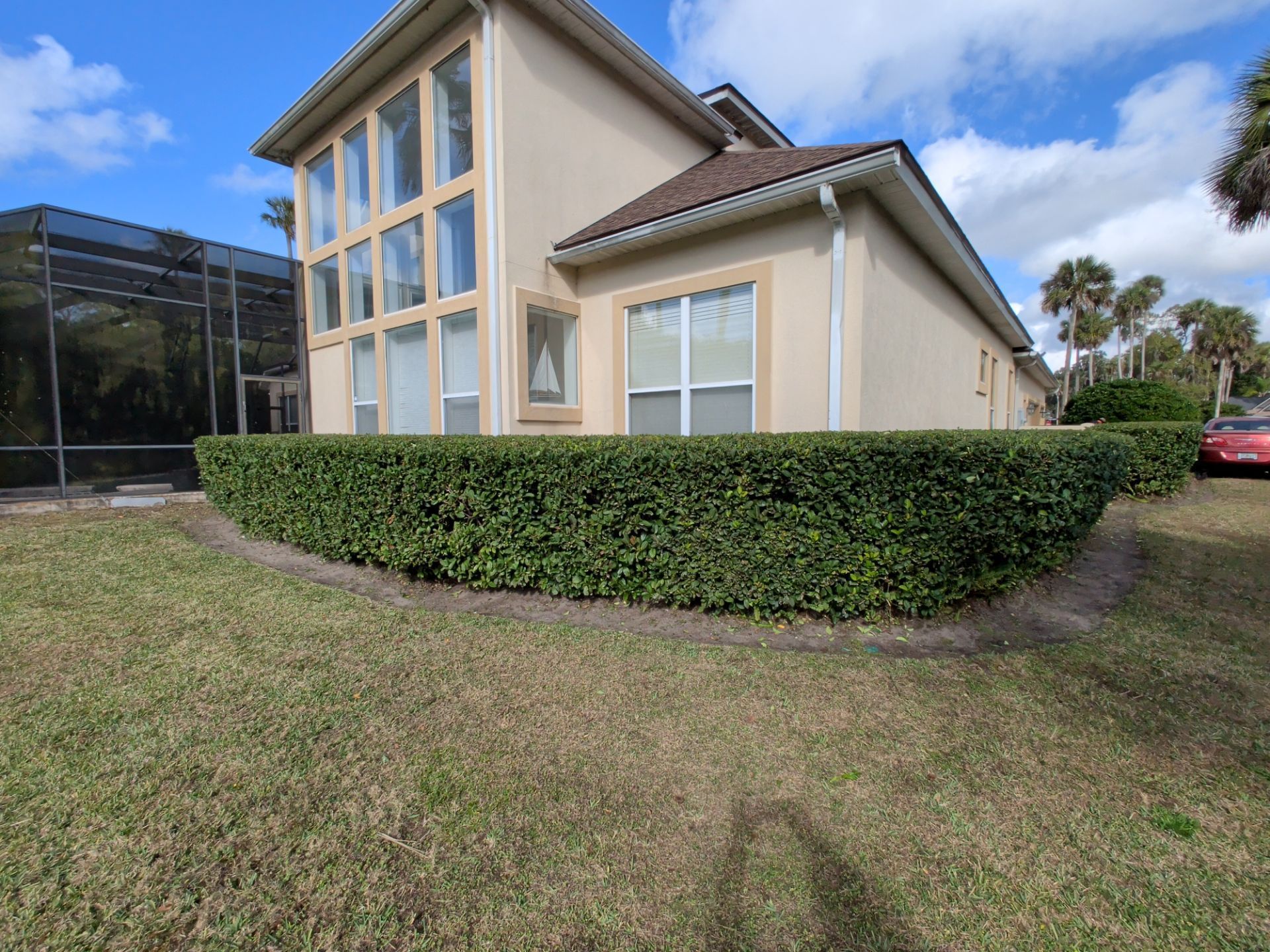Beige house with manicured green hedge and brown roof. Tall windows and a screened pool enclosure are visible.