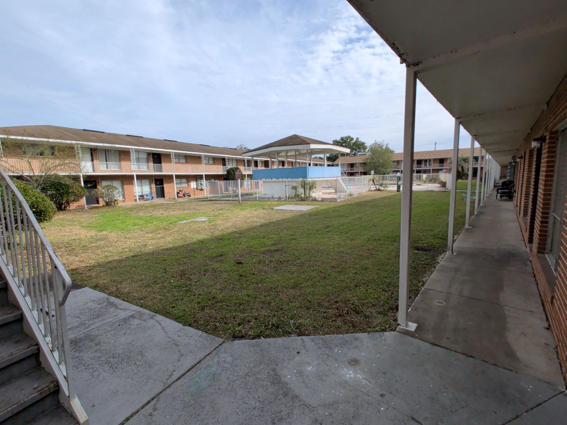 Courtyard of apartment buildings. Long covered walkway, lawn with sparse grass, a small pool area visible.