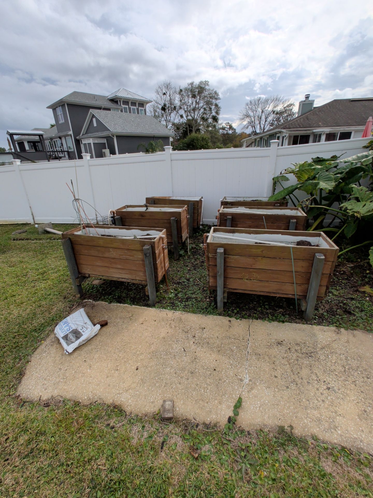 Six wooden raised garden beds in a backyard setting near a white fence and houses under an overcast sky.