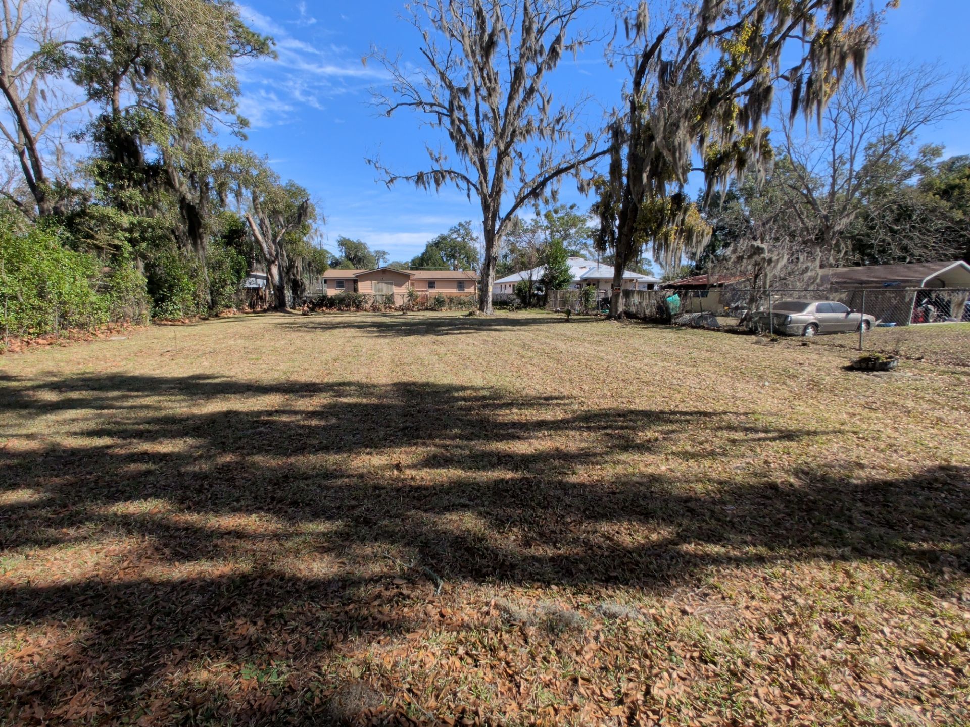 A vacant lot with brown grass, surrounded by trees and houses under a clear sky.