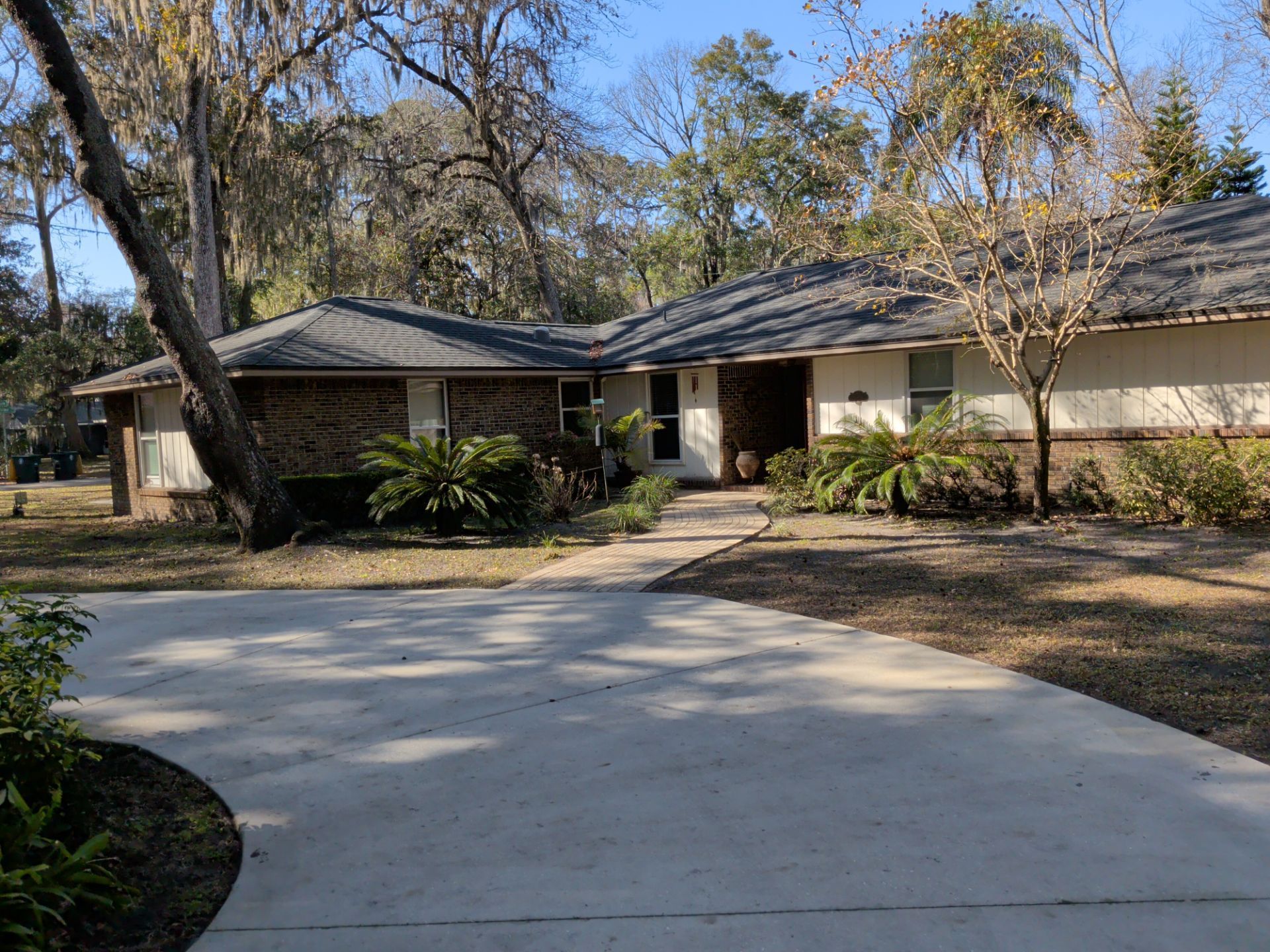 Ranch-style house with a curved concrete driveway. Brick and tan exterior, surrounded by trees and landscaping.