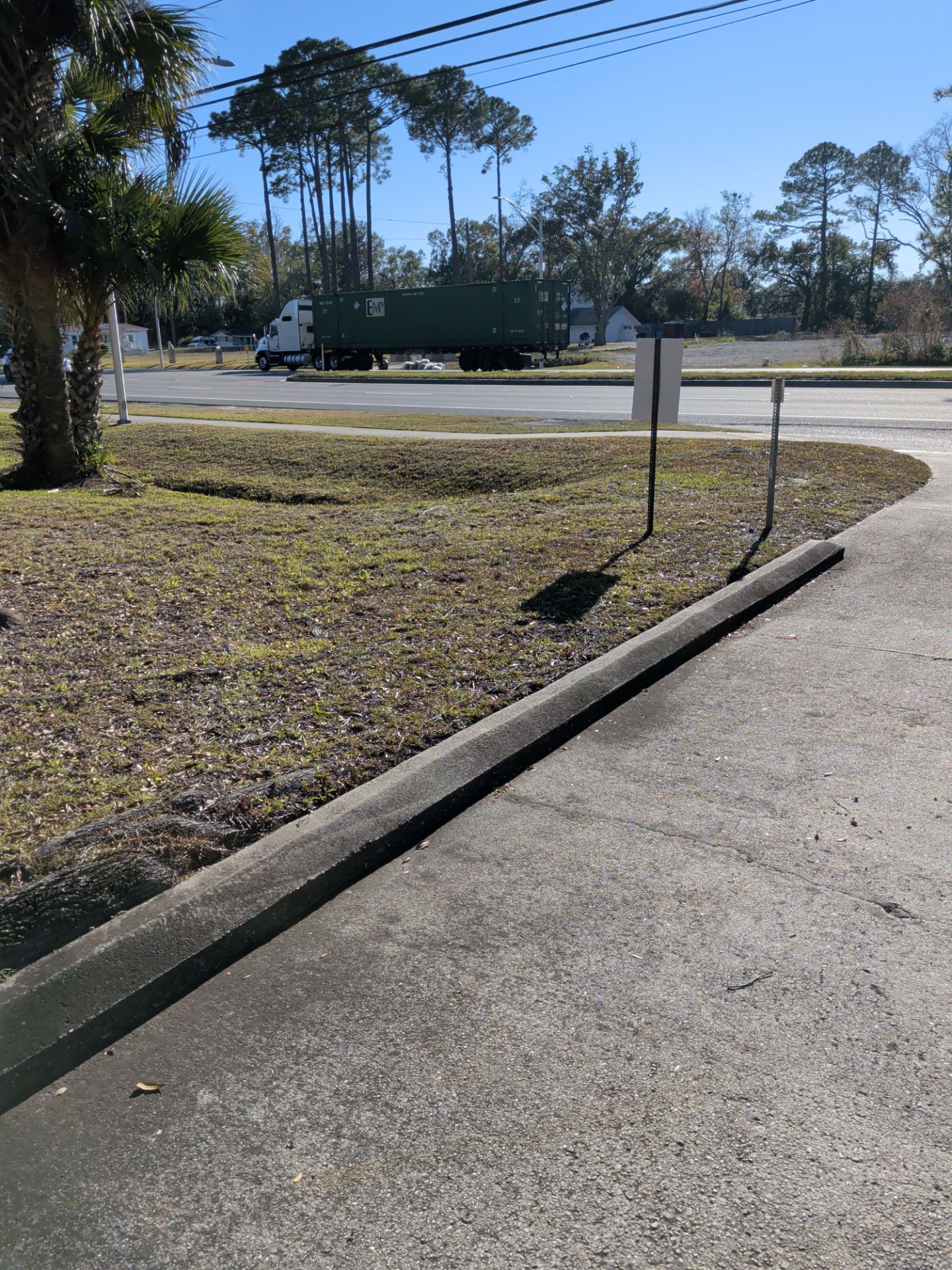 Concrete curb and grassy area with a truck visible in the distance. Blue sky and trees in the background.
