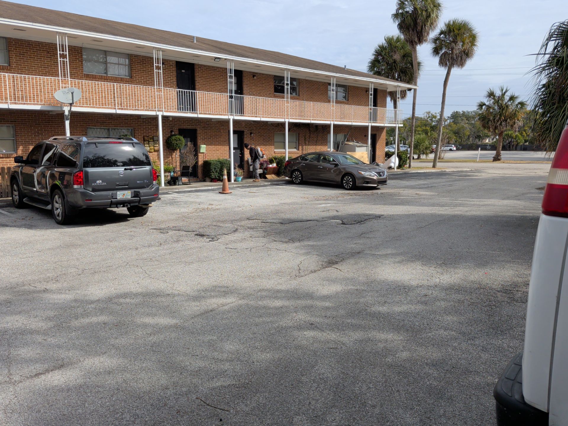 Two-story brick apartment building with cars parked in front on a gravel lot. Palm trees in background.