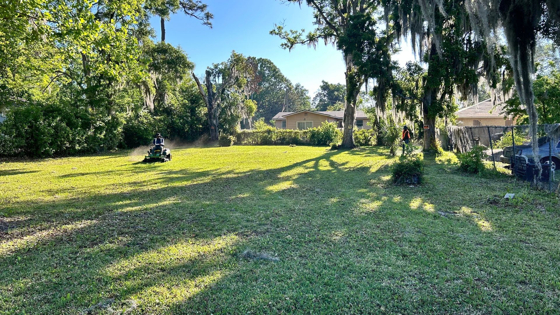 Man mowing lawn in a sunny yard with trees and houses in the background.