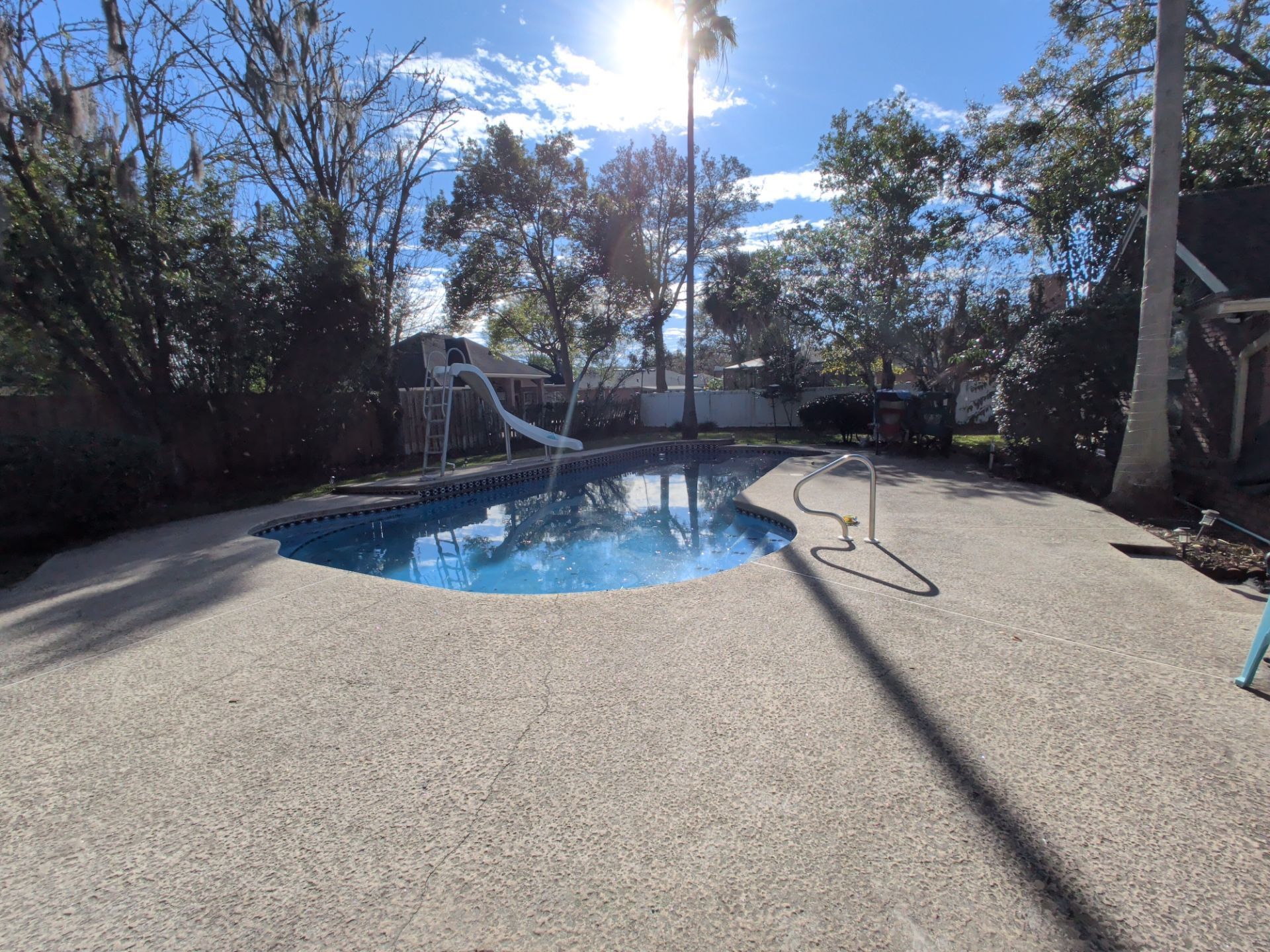 A backyard pool with a slide, surrounded by a concrete patio, trees, and a sunny sky.