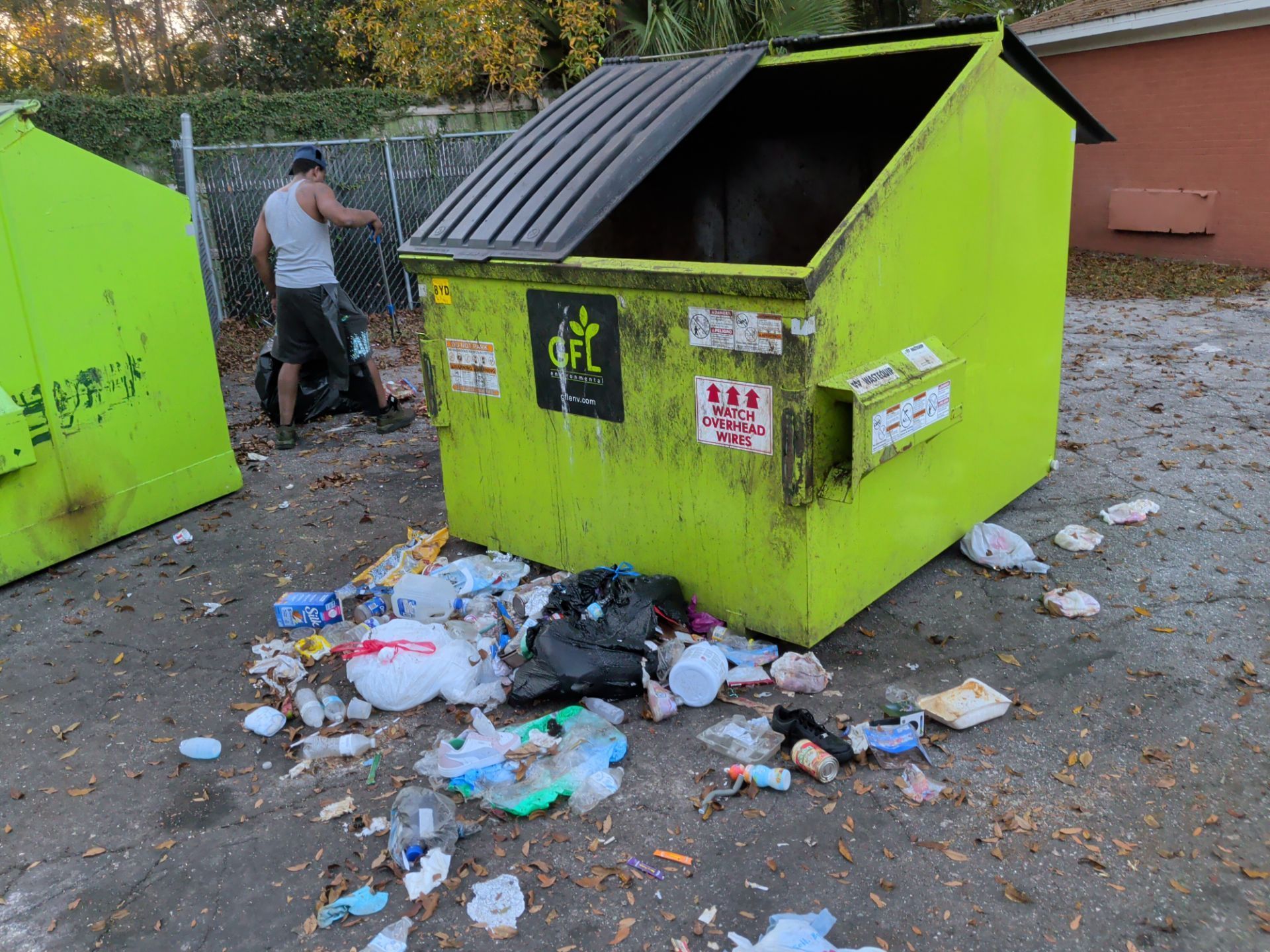 A person by a bright green dumpster with overflowing trash on a gray asphalt surface.