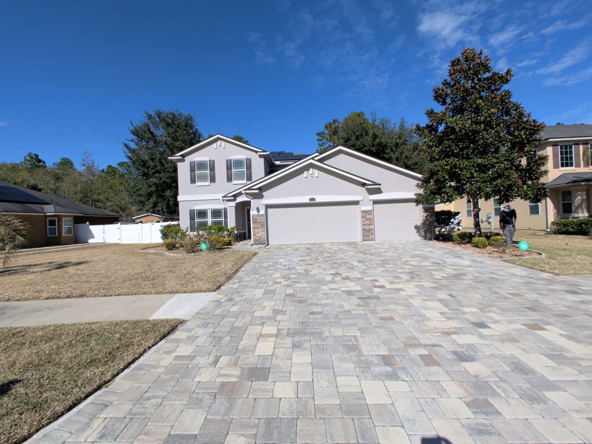 Two-story house with paver driveway on a sunny day. Gray exterior, two-car garage, and landscaped yard.