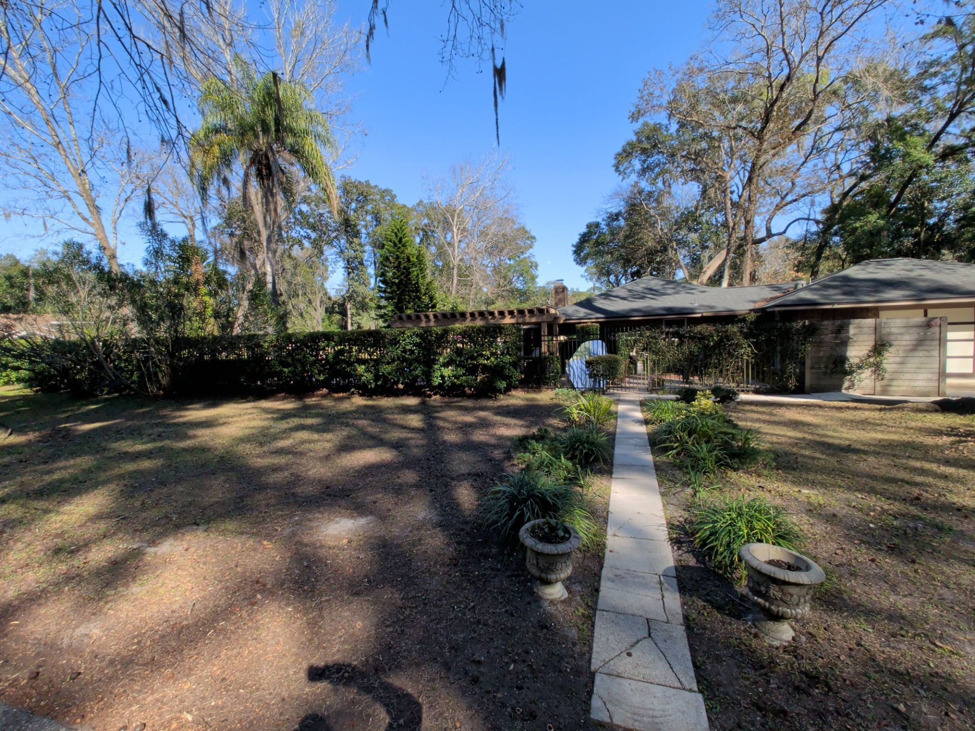 Stone path leads to a house with a pergola, flanked by grass, bushes, and trees on a sunny day.