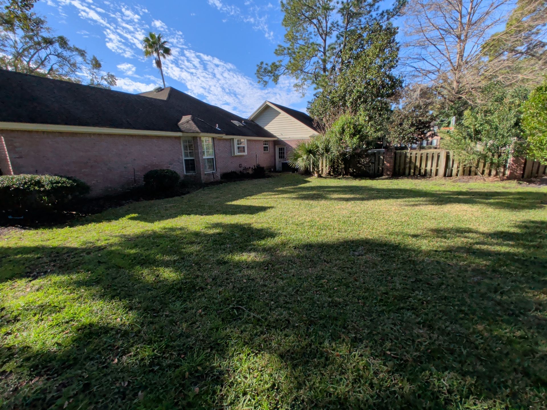 Backyard with brick house, green grass, trees, and wooden fence on a sunny day.