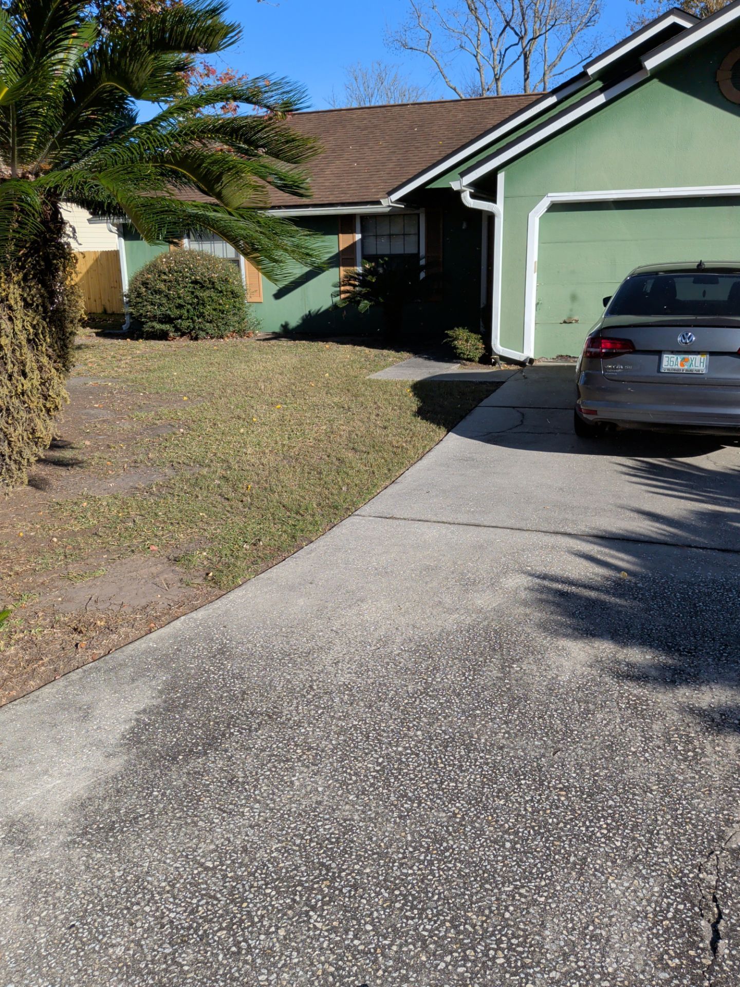 A house with green siding and brown roof. A gray car sits in the driveway. A patch of grass is visible.