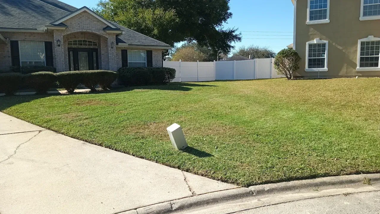 A house with patchy green lawn, a driveway, and a white fence in the background.