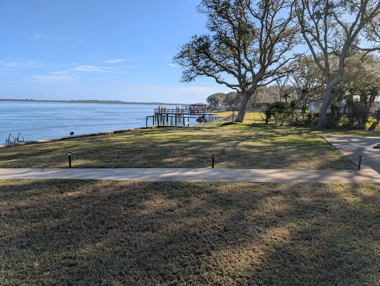 Waterfront scene: calm blue water, grassy shore, docks, trees, clear sky.