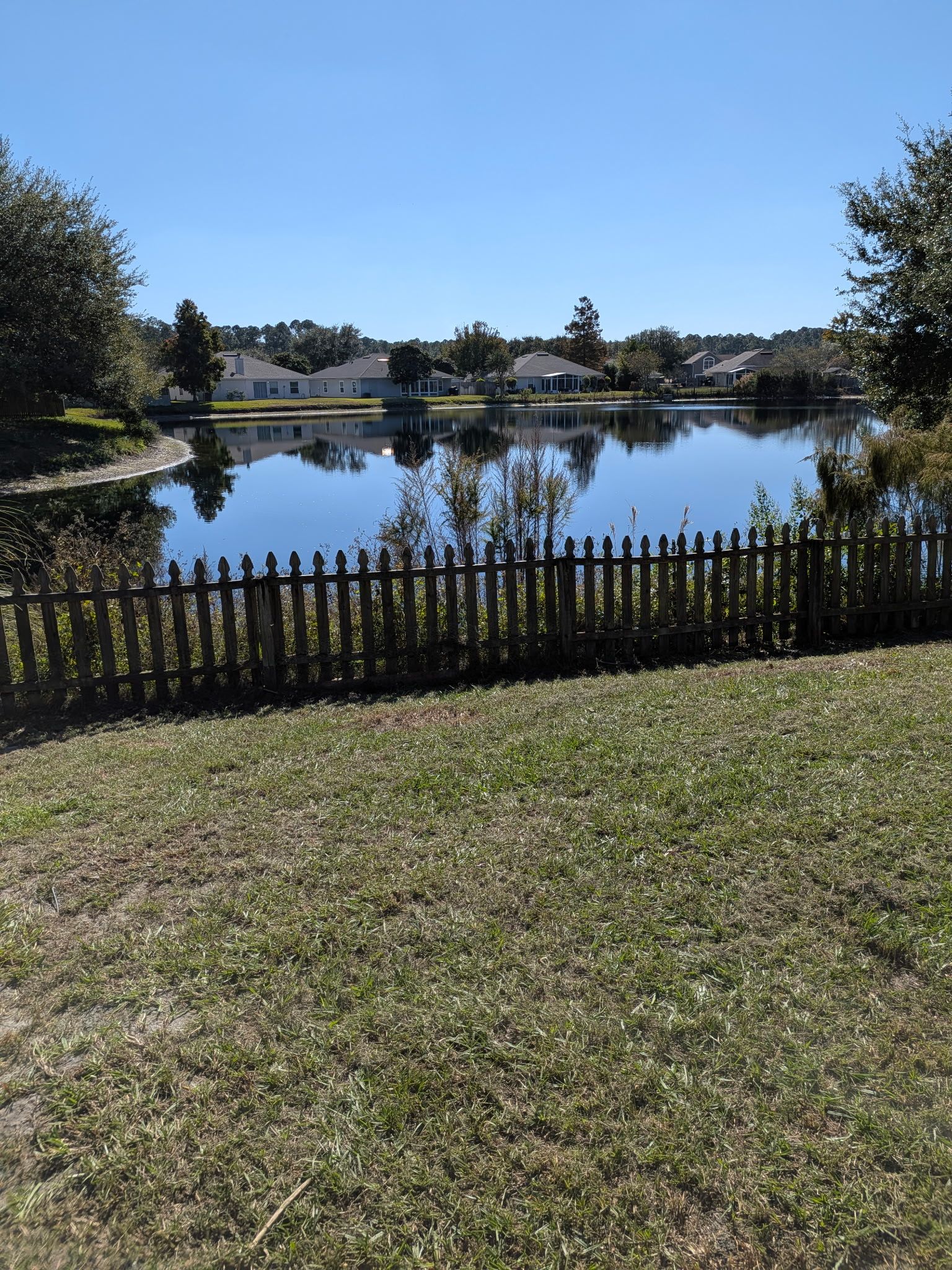 A calm lake reflects homes under a clear blue sky, viewed over a wooden fence and grassy lawn.