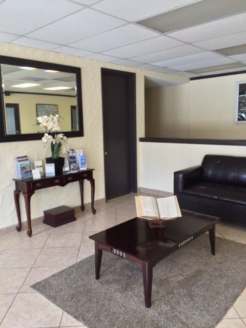 Waiting room with dark wood table, black sofa, side table, and wall mirrors on beige tile floor.