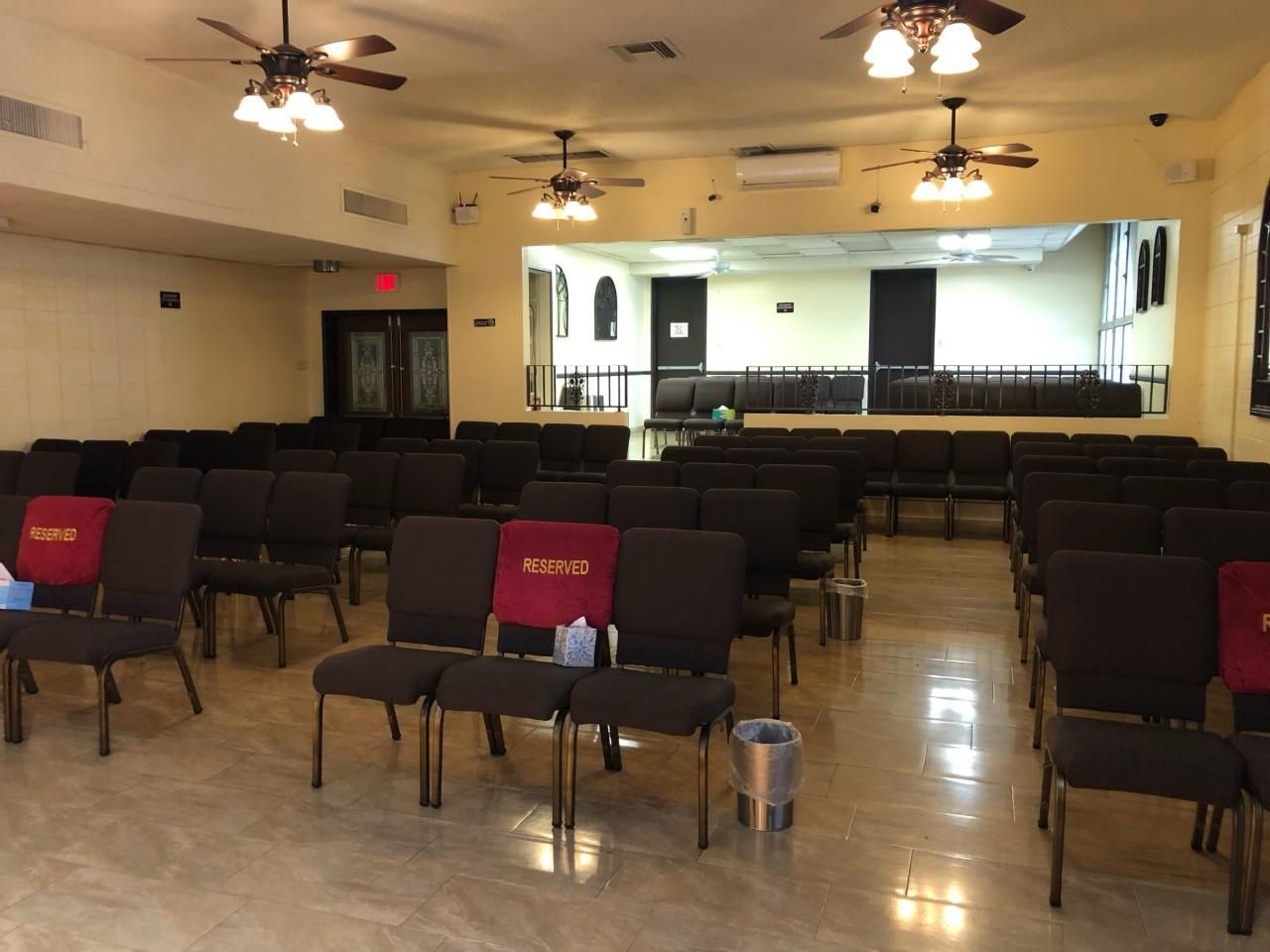 Empty meeting hall with rows of black chairs, ceiling fans, stage, and red EXIT signs