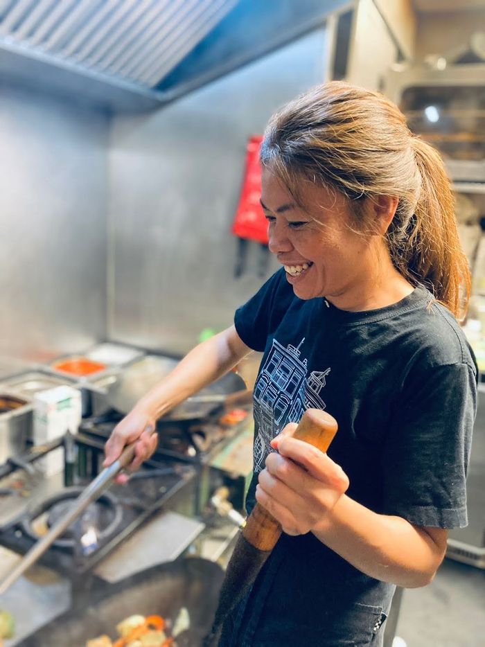 Een persoon in een zwart T-shirt lacht terwijl hij met een houten spatel in een metalen wok roert in een restaurantkeuken.
