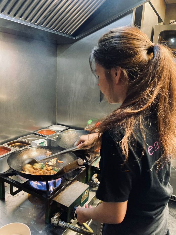 Een persoon in een zwart T-shirt roert voedsel in een wok boven een gasvlam in een professionele keuken van roestvrij staal.