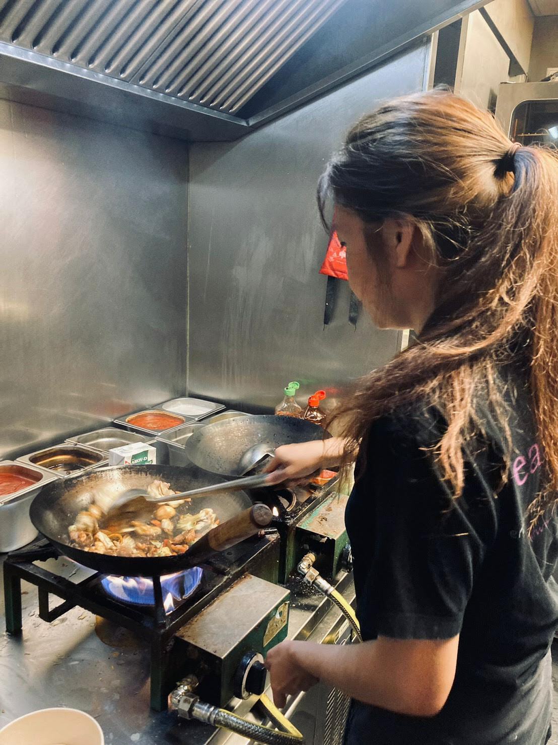 Een persoon in een zwart shirt bereidt voedsel in een wok boven een gasvlam in een professionele keuken van roestvrij staal.