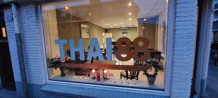 Een blik door een raam in restaurant Thai 88, met lichtblauwe en bruine letters en een interieur verlicht door kaarsen.