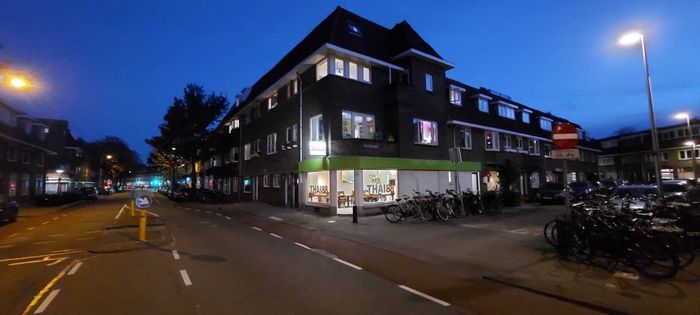 Een bakstenen hoekpand van meerdere verdiepingen in de schemering, met een verlichte etalage, geparkeerde fietsen en een straatlantaarn.