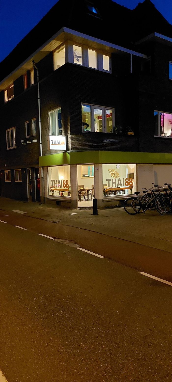 Een gebouw met meerdere verdiepingen in de schemering, met een fel verlicht Thais restaurant op de hoek van de begane grond.