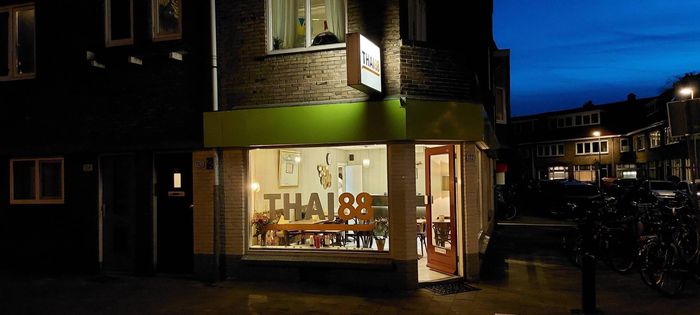 De gevel van een Thais restaurant (Thai 88) bij schemering, met een helder interieur in een donkere, meerlaagse stedelijke omgeving.