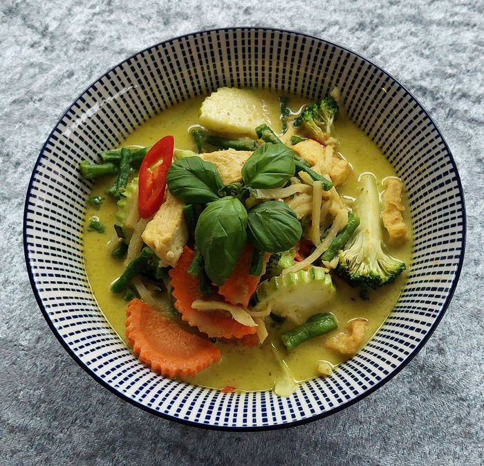 Een kom gele Thaise groene curry met tofu, broccoli, wortels en sperziebonen, gegarneerd met verse basilicum en chilipeper.